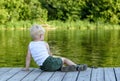 Little blond boy sitting on a pier on the bank of the river amid a green forest Royalty Free Stock Photo