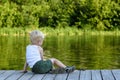 Little blond boy sitting on a pier on the bank of the river amid a green forest Royalty Free Stock Photo