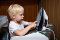 Little blond boy touches electric iron with his finger. Home safety