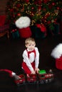 Little blond boy in white shirt and red bow tie with suspenders playing with a toy red steam train lying on floor in living room. Royalty Free Stock Photo