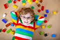 Little blond child with glasses playing with lots of colorful plastic blocks indoor. Kid boy wearing colorful shirt and Royalty Free Stock Photo