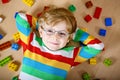 Little blond child with glasses playing with lots of colorful plastic blocks indoor. Kid boy wearing colorful shirt and Royalty Free Stock Photo