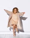 Little blonde model in golden necklace, fluffy pink dress and fur coat, shoes. Spread her hands, smiling, posing on white