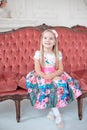 A little blond girl in colorful dress sitting on big sofa