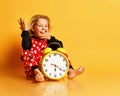 Little blond girl red dotted home clothing sitting on floor with big yellow alarm clock and yawning over yellow background Royalty Free Stock Photo