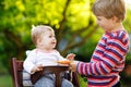 Little blond kid boy giving a carrot to baby sister. Happy siblings having healthy snack. Baby girl sitting in high