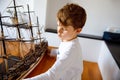 Little blond school kid boy playing with sailing ship model indoors. Excited child with yacht having fun after school at Royalty Free Stock Photo