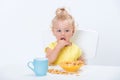 Little blonde baby girl 1 year old in yellow t-shirt eating cereal flakes and drinking a cup of milk tea at the table isolated on Royalty Free Stock Photo