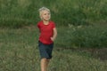 Little blonde boy in red t-shirt walks on green field. Happy running boy. Childhood Royalty Free Stock Photo