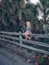 Little blonde Caucasian girl in pink skirt sitting on wooden fence relaxing. Child dreaming among tall palm trees on summer day Royalty Free Stock Photo