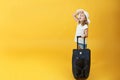 A little blonde child girl is ready to travel, stands with a suitcase and looks into the sky. Yellow background Royalty Free Stock Photo