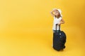 A little blonde child girl is ready to travel, stands with a suitcase and looks into the sky. Yellow background Royalty Free Stock Photo