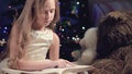 A little blonde girl in a festive dress with a book in her hands sits next to soft toys against the background of a Royalty Free Stock Photo