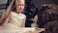 A little blonde girl in a festive dress with a book in her hands sits next to soft toys against the background of a Royalty Free Stock Photo