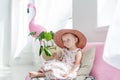 Little blonde girl in hat sitting on a couch in her room. Small girl and pink flamingo indoors Royalty Free Stock Photo