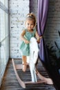 A little blonde girl is having fun and swinging on a wooden rocking horse near the window Royalty Free Stock Photo