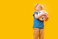 Little blonde girl hugs soccer ball. yellow studio background. Copy space, mockup. Football for kids Royalty Free Stock Photo