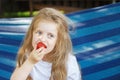 Little blonde girl with long hair eats a strawberry in the garden sitting in a blue hammock Royalty Free Stock Photo