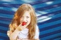 little blonde girl with long hair eats a strawberry in the garden sitting in a blue hammock Royalty Free Stock Photo