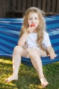 Little blonde girl with long hair eats a strawberry in the garden sitting in a blue hammock Royalty Free Stock Photo