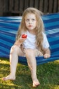 little blonde girl with long hair eats a strawberry in the garden sitting in a blue hammock Royalty Free Stock Photo