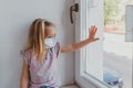 A little blonde girl looking out the window of the face mask from coronavirus. quarantine in Europe. stay home covid Royalty Free Stock Photo