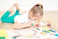 Little blonde girl painting on big white paper while laying on the floor indoors