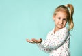 Little blonde girl with ponytail, in shirt with hearts print. She smiling, holding something posing on blue background. Close up Royalty Free Stock Photo