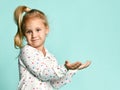 Little blonde girl with ponytail, in shirt with hearts print. She smiling, holding something posing on blue background. Close up Royalty Free Stock Photo