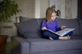 a little blonde girl is reading a big book sitting on the sofa in the living room.Smile and interest in reading.Self Royalty Free Stock Photo