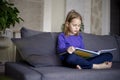 a little blonde girl is reading a big book sitting on the sofa in the living room.Smile and interest in reading.Self Royalty Free Stock Photo