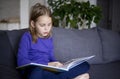 a little blonde girl is reading a big book sitting on the sofa in the living room.Smile and interest in reading.Self Royalty Free Stock Photo