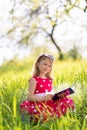 Little blonde girl in a red dress reads a book with fairy tales in nature Royalty Free Stock Photo