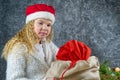 Little blonde girl in a red gnome hat unhappy with a bag of gifts from Santa in anticipation of a Christmas miracle