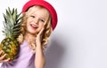 Little blonde girl in red hat and purple blouse. She is smiling, touching her face and holding pineapple, posing  on white Royalty Free Stock Photo