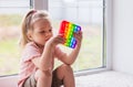 A little blonde girl sits near the window and plays with new trend sensory toy - rainbow pop it