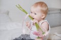 Little blonde girl sitting on the bed in a light top and gray pants and biting flower stalks, pink gerberas Royalty Free Stock Photo