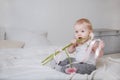 Little blonde girl sitting on a bed with pink flowers and a ribbon and biting them, portrait of a cute baby on a light background Royalty Free Stock Photo