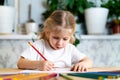 a little blonde girl is sitting at the table, smiling and drawing a heart with a red pencil, there are a lot of colored Royalty Free Stock Photo