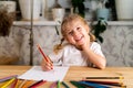 A little blonde girl is sitting at the table, smiling and drawing a heart with a red pencil, there are a lot of colored pencils on Royalty Free Stock Photo