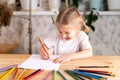 a little blonde girl is sitting at the table, smiling and drawing a heart with a red pencil, there are a lot of colored Royalty Free Stock Photo