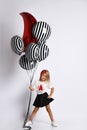 Little blonde girl in t-shirt, red headband, black skirt and sneakers. She holding balloons, posing isolated on white. Full length Royalty Free Stock Photo