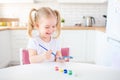 Little blonde girl with two ponytales playing and painting on her hands with blue paint sitting at the light kitchen at home. Self Royalty Free Stock Photo