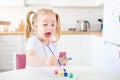 Little blonde girl with two ponytales playing and painting on her hands with blue paint sitting at the light kitchen at home. Self Royalty Free Stock Photo