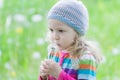 Little blonde girl wearing striped knitted hat blowing on white puffy dandelion seed head at her hand