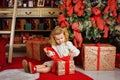 A little blonde girl in a white dress opens a gift sitting near the Christmas tree and lights of garlands Royalty Free Stock Photo