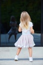 A little blonde girl in a white t-shirt and pink skirt looks at her reflection in the glass surface Royalty Free Stock Photo