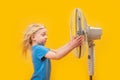 Little blonde girl with windblown hair near fan on yellow background. Fair-haired child near blower