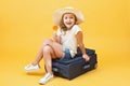 A little blonde kid girl dreams of traveling and sits on a suitcase with candy in her hand. Yellow background