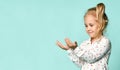 Little blonde kid with ponytail, in shirt with hearts print. She smiling, holding something posing on blue background. Close up Royalty Free Stock Photo